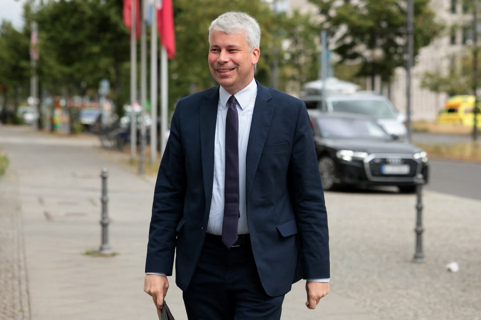 Steffen Bilger, smiling, walks along a wide pavement in a dark navy suit, light blue shirt and dark tie, holding a folder; flagpoles, trees and a parked dark car behind him