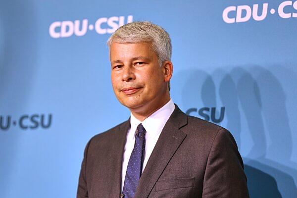 Steffen Bilger in a brown suit, white shirt and dotted blue tie, standing in front of a light blue backdrop printed with the CDU/CSU logo