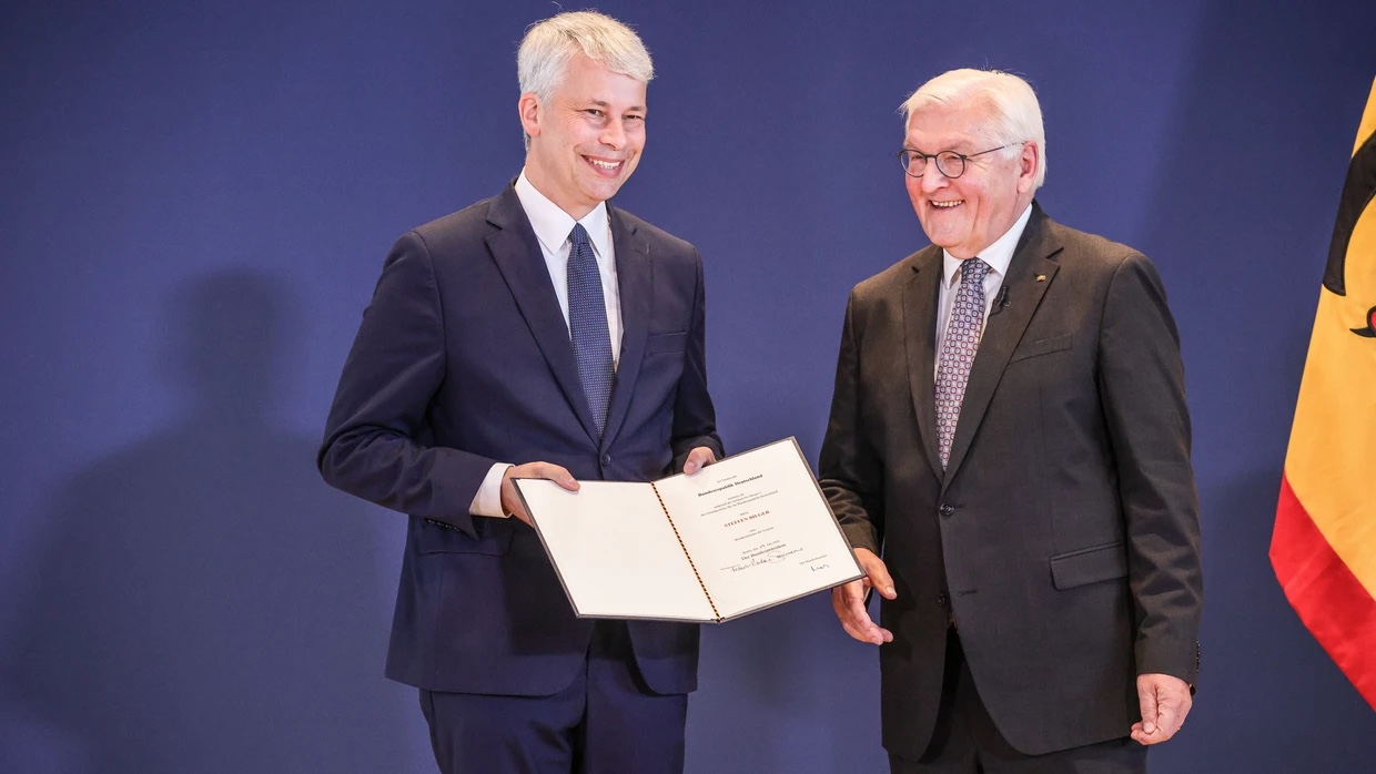 Steffen Bilger, smiling, holds open his certificate of appointment next to Federal President Frank-Walter Steinmeier in front of a blue wall and the German federal flag