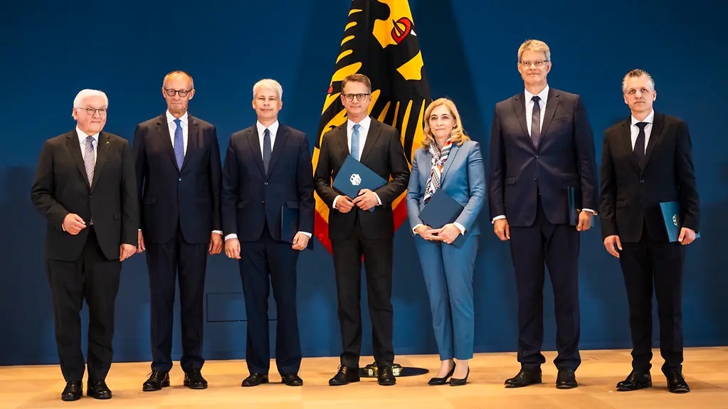 Federal President Frank-Walter Steinmeier standing in a row with six other people, several holding blue certificate folders, in front of the German federal flag