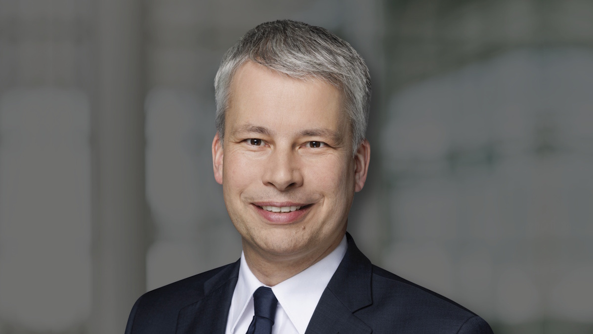 Portrait of Steffen Bilger in a dark suit and white shirt, smiling, against a softly blurred grey background