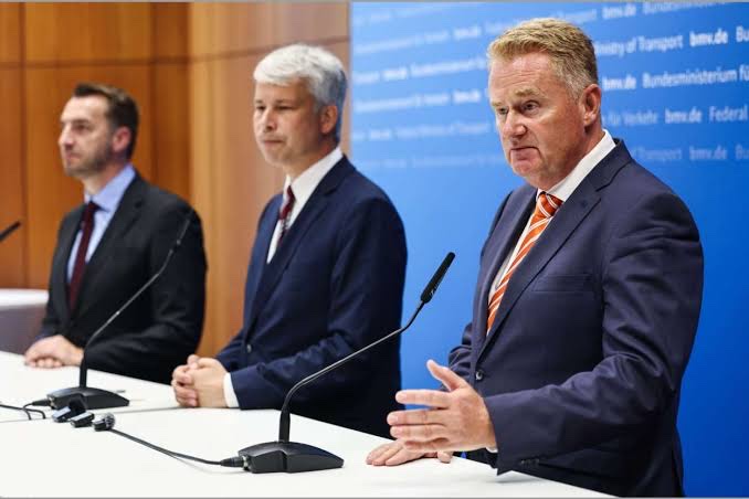 Three men in suits at a white podium table with microphones in front of a blue Federal Ministry for Transport backdrop; Steffen Bilger sits in the middle while the man on the right, in an orange tie, speaks