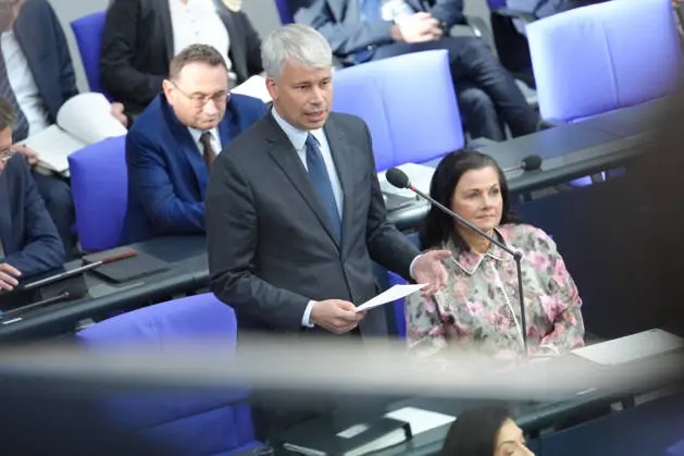 Steffen Bilger standing in the plenary chamber among seated members of parliament, reading from a sheet of paper