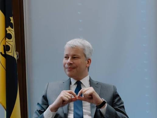 Steffen Bilger in a grey suit and blue tie, smiling and forming a heart shape with his hands, with the black-and-gold flag of Baden-Württemberg on the left