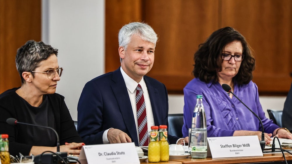 Steffen Bilger, smiling, seated at a conference table between two women, with a name card reading 'Steffen Bilger MdB, Bundesminister, BMV', microphones and bottles of juice and water in front of him