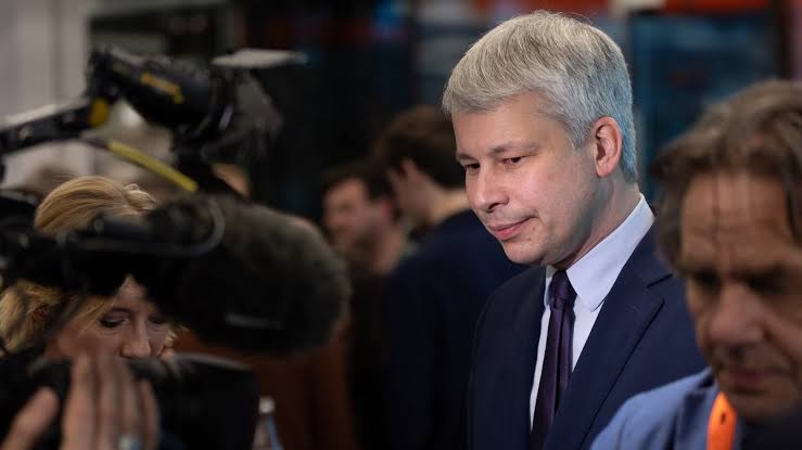 Steffen Bilger, in a dark suit and purple tie, looking down thoughtfully while surrounded by television camera crews and a boom microphone