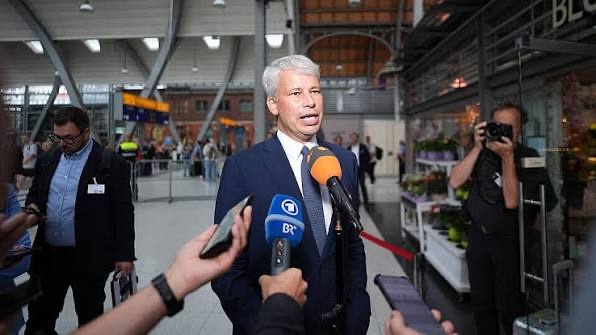 Steffen Bilger speaks into a cluster of television microphones in a large railway station concourse, with travellers, a photographer and departure boards behind him