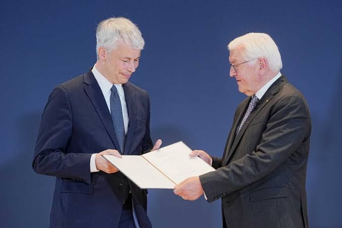 Federal President Frank-Walter Steinmeier hands Steffen Bilger an open certificate of appointment in a white folder in front of a dark blue backdrop