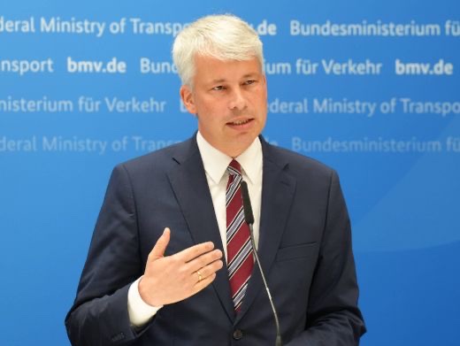 Steffen Bilger in a dark suit and red striped tie speaking at a lectern microphone in front of a blue Federal Ministry for Transport backdrop reading bmv.de