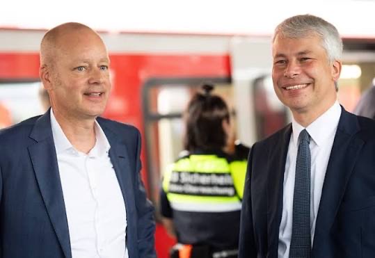 Steffen Bilger, smiling, stands beside a bald man in a dark jacket in front of a red train; behind them a security staff member in a high-visibility vest marked Sicherheit faces the train